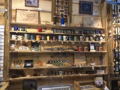 A wooden shelf displays various mugs, artwork, and crafts, creating a cozy, rustic shop atmosphere.