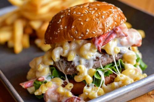 A close-up of a cheeseburger topped with macaroni and cheese, served with a side of fries on a metal tray.