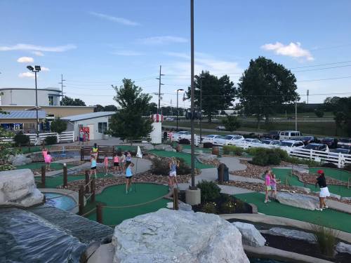 A mini-golf course with children playing, surrounded by greenery and rocks, under a clear blue sky.