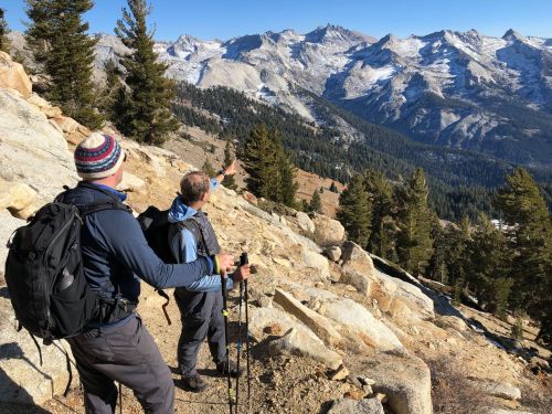 Two hikers on a rocky trail, pointing towards a distant mountain range surrounded by trees under a clear blue sky.