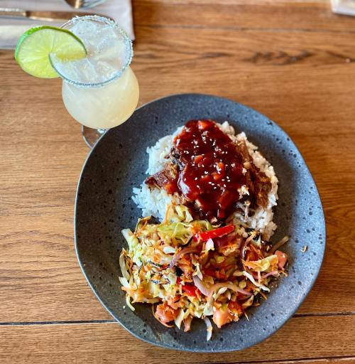 A plate of rice topped with meat and sauce, accompanied by a colorful cabbage salad and a cocktail with a lime garnish.