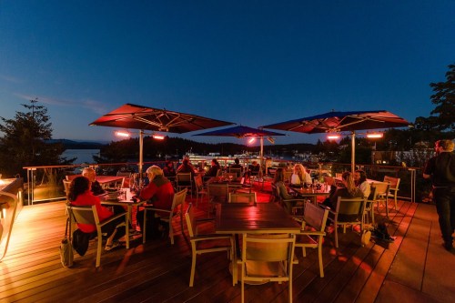 Outdoor dining area at dusk, featuring tables under umbrellas with heaters, overlooking a scenic view.