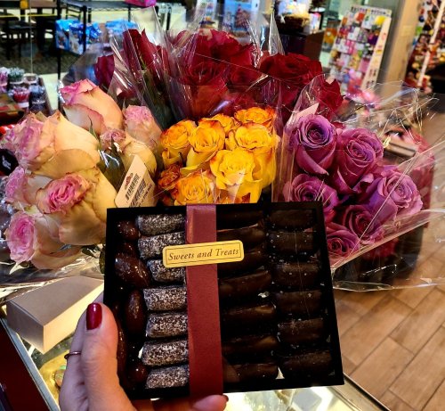 A hand holds a box of chocolates labeled "Sweets and Treats" in front of colorful flower bouquets.