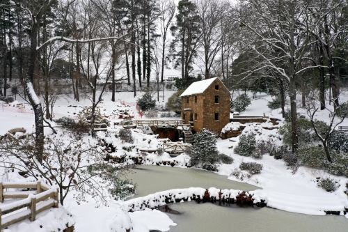 A snowy landscape featuring a stone building by a frozen pond, surrounded by trees and snow-covered ground.