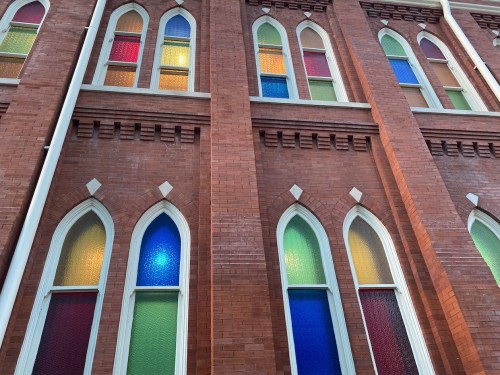 Colorful stained glass windows in a brick building, featuring arched designs in red, blue, green, and yellow hues.