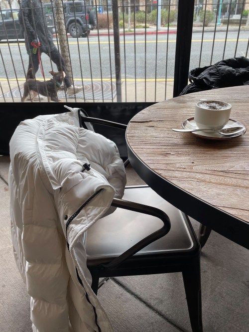 A white puffer jacket draped over a chair next to a table with a cup of coffee, with a street view in the background.