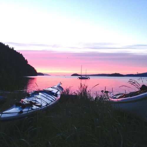 A serene sunset over calm waters, with kayaks on the shore and silhouettes of distant islands.