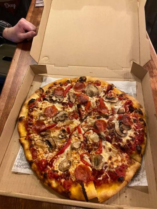 A freshly baked pizza with pepperoni, mushrooms, and bell peppers in an open pizza box on a wooden table.