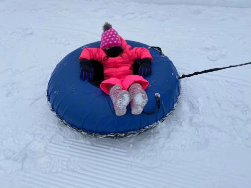 A child in a pink snowsuit sits in a blue snow tube on a snowy surface, ready for tubing fun.