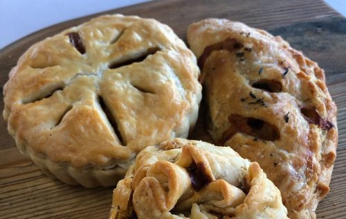 Three freshly baked pastries on a wooden board, including a round pie and two hand pies with flaky crusts.