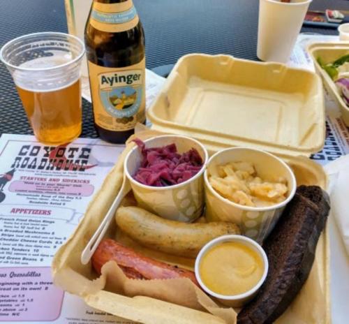 A takeout container with a sausage, macaroni and cheese, pickled red cabbage, and mustard, alongside a beer and menu.