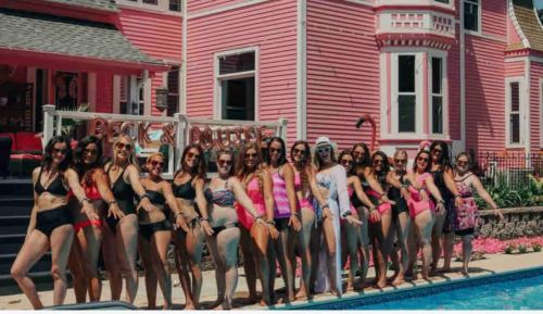 A group of women in swimsuits poses by a pink house and pool, enjoying a sunny day.