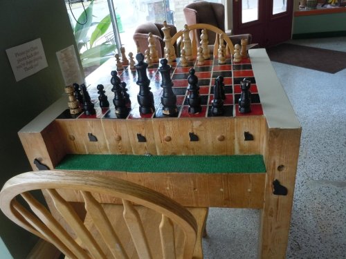 A large, wooden chess table with black and white pieces, set in a cozy indoor space with a chair nearby.