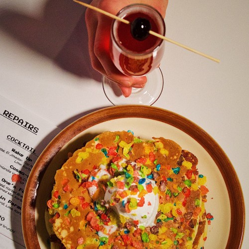 A hand holds a cocktail above a plate of colorful pancakes topped with whipped cream and cereal.