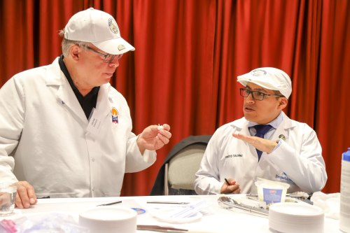 Two men in white lab coats and hats discuss over a table with scientific equipment and materials.
