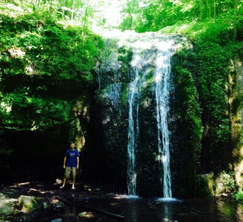 A person stands near a waterfall surrounded by lush greenery and sunlight filtering through the trees.