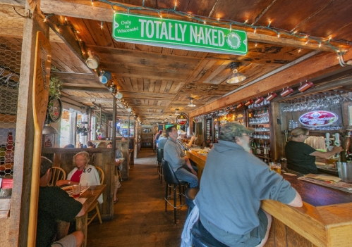 A rustic bar interior with wooden decor, patrons at the counter, and a sign reading "Totally Naked" hanging above.