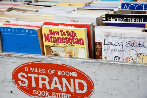 A wooden crate filled with various books, featuring titles like "How to Talk Minnesotan" and "Loving Edith."