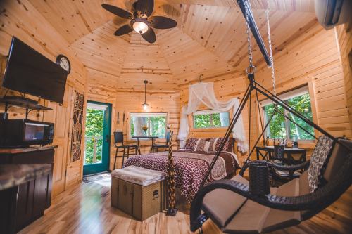 Cozy wooden cabin interior with a bed, swing chairs, and large windows overlooking greenery. Warm, inviting atmosphere.