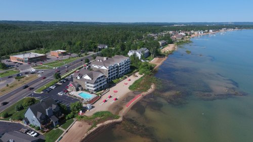 Aerial view of a lakeside resort with sandy beach, green trees, and nearby buildings along the shore. Clear blue sky.
