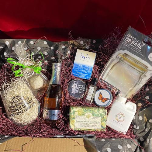 A gift box filled with various items, including a bottle, candles, soap, and a mason jar bag, arranged with decorative filler.