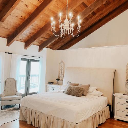 Cozy bedroom with a large bed, chandelier, and wooden beams, featuring a chair and sliding glass doors.