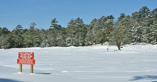 A snowy landscape with a red safety sign in the foreground and trees in the background under a clear blue sky.