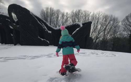A child in colorful winter clothing walks through snow, with a large, abstract structure and trees in the background.