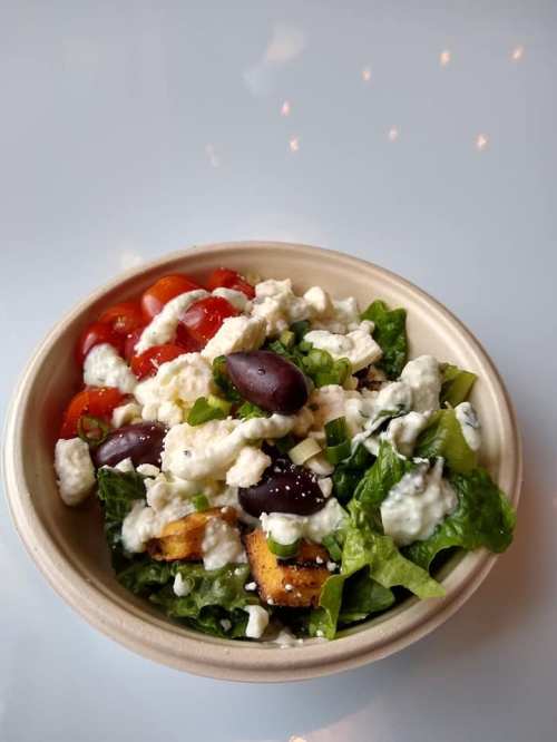 A bowl of salad with lettuce, cherry tomatoes, olives, croutons, and feta cheese on a light background.