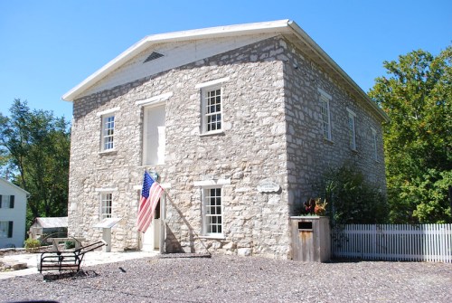 Historic stone building with large windows, an American flag, and a white picket fence under a clear blue sky.