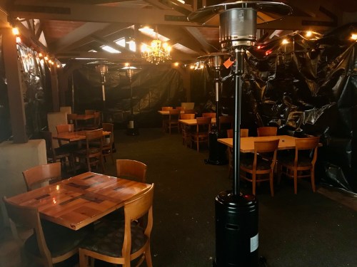 Cozy indoor dining area with wooden tables, chairs, and patio heaters, surrounded by black plastic sheeting.