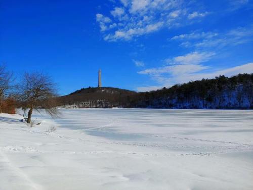 A snowy landscape with a frozen lake and a tall monument on a hill under a clear blue sky.