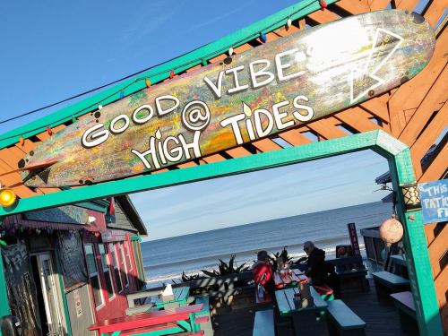 A colorful sign reading "Good Vibes @ High Tides" above a beachside deck with ocean views and people sitting below.