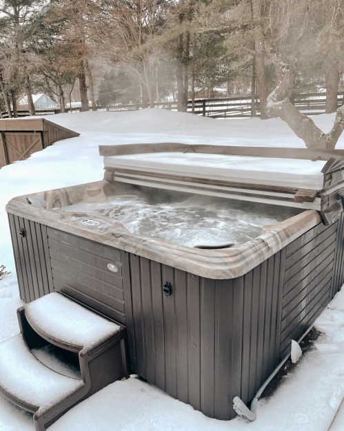 A hot tub with steam rising, surrounded by snow and trees, creating a cozy winter scene.