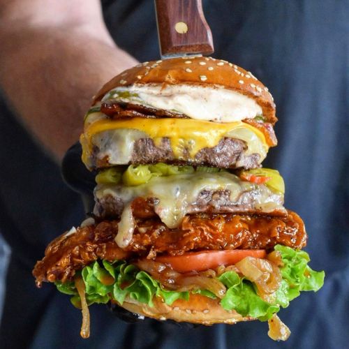 A towering burger with multiple patties, cheese, lettuce, tomato, and a knife through the top, held by a person.
