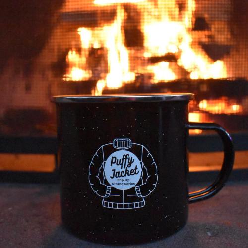 A black mug with a "Puffy Jacket" logo in front of a warm, glowing fireplace.