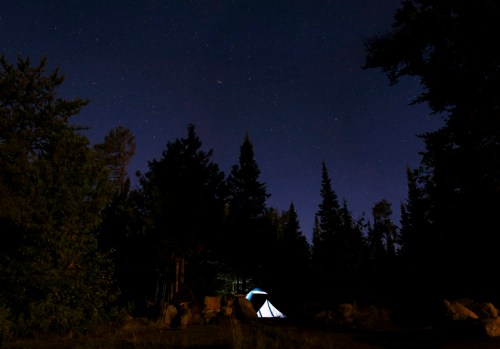 A tent illuminated under a starry night sky, surrounded by tall trees and rocky terrain.