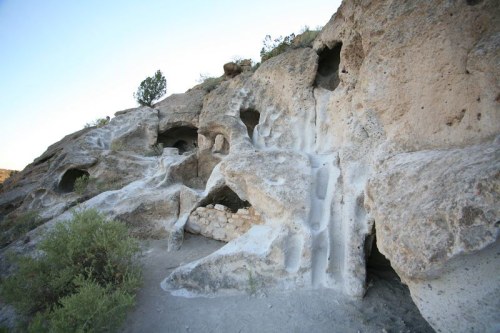 Rocky cliffside with natural caves and erosion patterns, surrounded by sparse vegetation and clear blue sky.