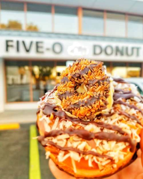 A close-up of a donut topped with chocolate, coconut, and a cookie, with a donut shop in the background.