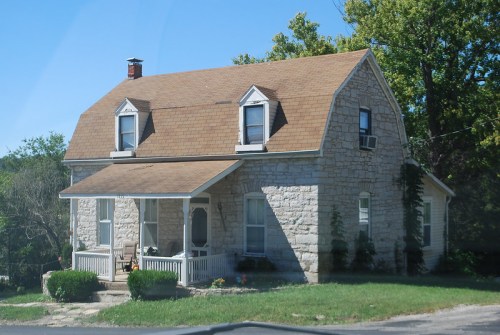 A charming stone house with a brown roof, surrounded by greenery and a small porch, set against a clear blue sky.