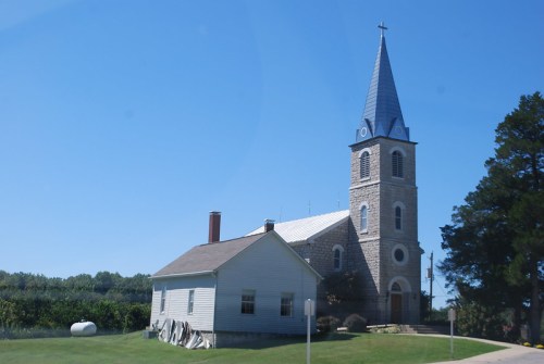 A stone church with a tall steeple and a nearby white building, set against a clear blue sky.