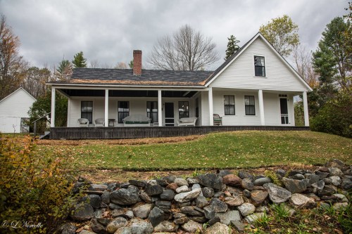 A white farmhouse with a porch, surrounded by trees and a stone wall, under a cloudy sky.