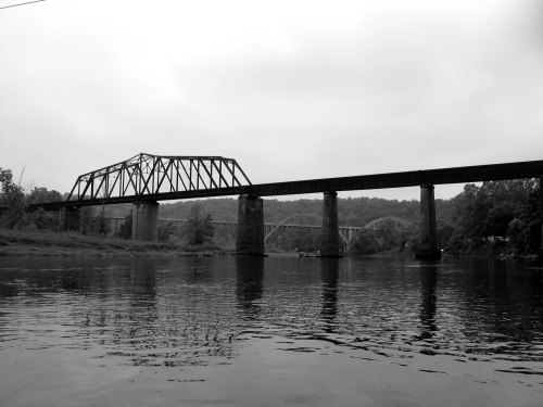 Black and white image of two bridges spanning a river, surrounded by trees under a cloudy sky.