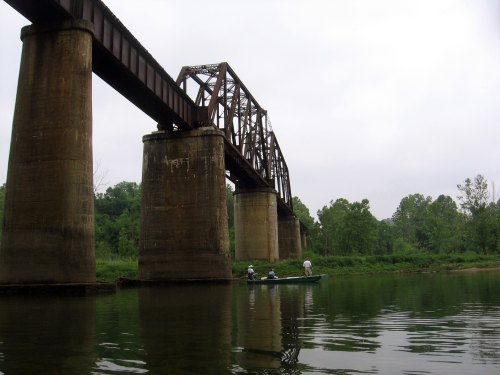 A boat with two people floats on a river beneath a large, rusted railway bridge surrounded by greenery.