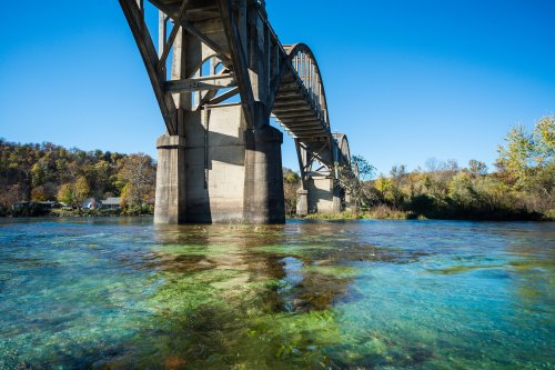 A clear river flows beneath a large, arched bridge, surrounded by trees and a bright blue sky.
