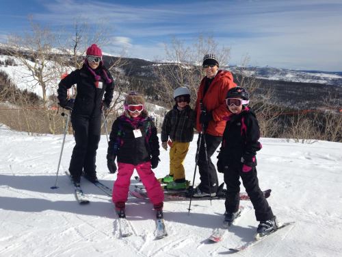 A group of five people, including children, dressed in ski gear, posing on a snowy slope with mountains in the background.