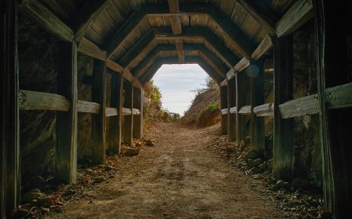 A dimly lit tunnel with wooden supports, leading to a bright opening at the end, surrounded by rocky terrain.