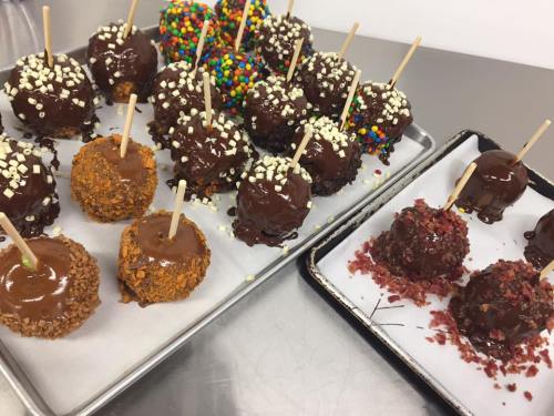 A tray of chocolate-covered treats on sticks, topped with colorful sprinkles and nuts, arranged neatly on parchment paper.
