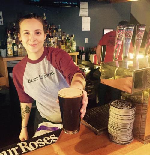 A bartender smiles while holding a pint of dark beer at a bar, with shelves of liquor in the background.