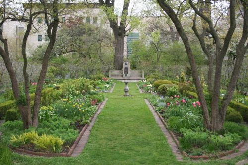 A lush garden with vibrant flowers, neatly trimmed hedges, and a central fountain surrounded by trees.